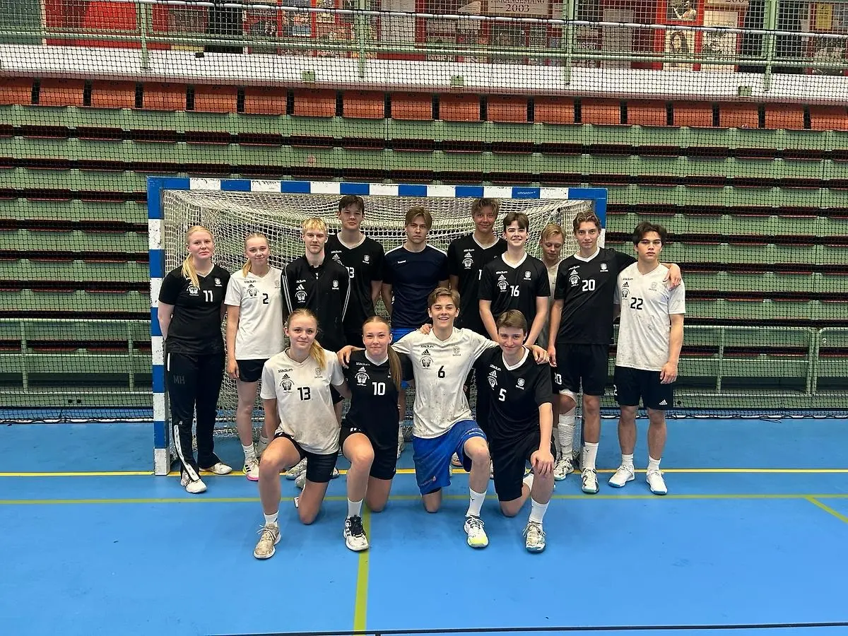 Gruppbild på ett handbollslag bestående av ungdomar, både killar och tjejer, som poserar framför ett mål inne i en idrottshall. Några spelare står upp medan andra knäar framför dem. Alla är klädda i matchställ i svart eller vitt.